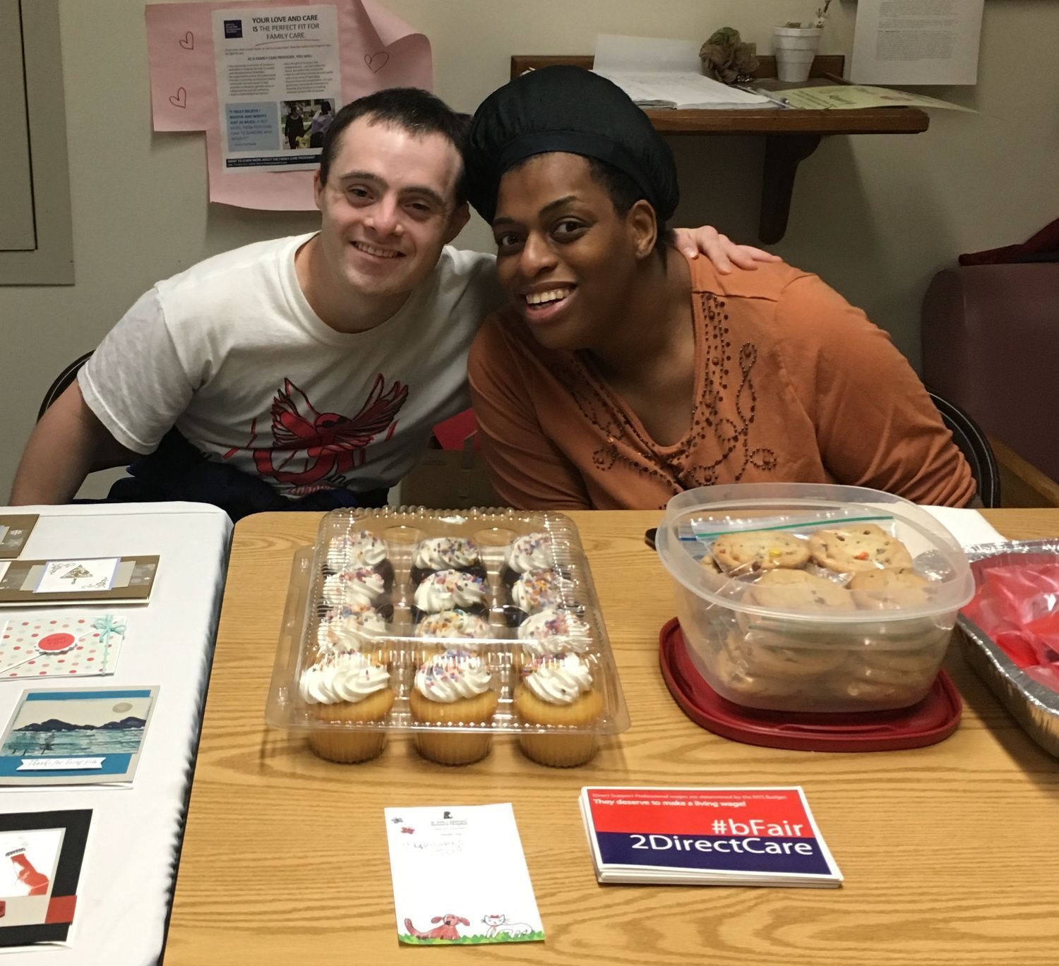 a white man with down syndrome and a black woman with physical disabilities lean in together over a table of cupcakes